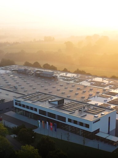 Aerial view of a modern building with a flat roof, surrounded by greenery and foggy landscapes during sunrise.