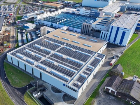 Luftaufnahme einer modernen Fabrikanlage mit Solarzellen auf dem Dach, umgeben von Parkplätzen und Grünflächen.