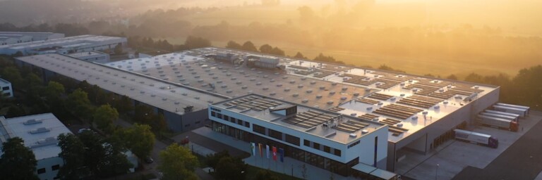 Luftaufnahme eines modernen Industriegebäudes mit flachem Dach und Solarpanels, umgeben von Bäumen und einer ländlichen Landschaft im Hintergrund.