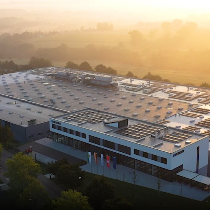 Luftaufnahme eines modernen Industriegebäudes mit flachem Dach und Solarpanels, umgeben von Bäumen und einer ländlichen Landschaft im Hintergrund.