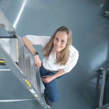 A woman standing on a ladder and smiling, wearing a white shirt and blue jeans, with a gray floor in the background.