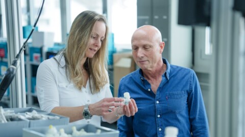 A man and a woman examining a small object together in a bright, modern workspace filled with boxes and equipment.