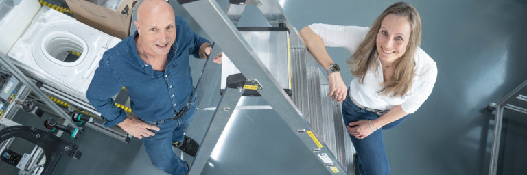 Two people smiling on a metal ladder in a workspace, with tools and materials in the background.