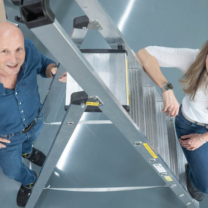 Two people smiling on a metal ladder in a workspace, with tools and materials in the background.