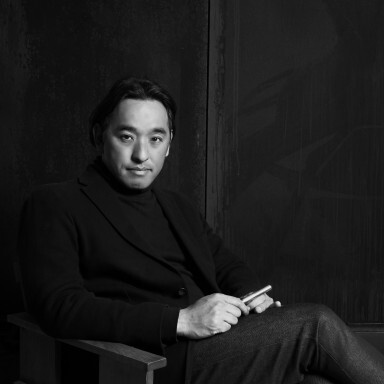 A man seated on a wooden chair, wearing a turtleneck and blazer, with a serious expression in a monochrome setting.