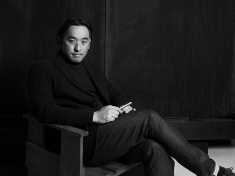 A man seated in a wooden chair, wearing a black turtleneck and blazer. The image is black and white.