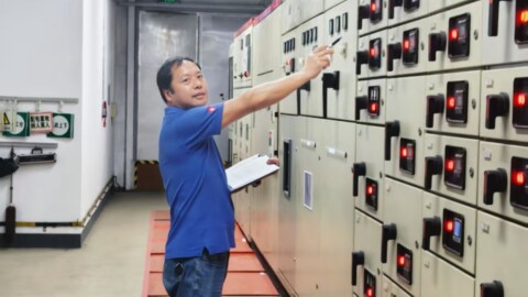 A technician in a blue shirt pointing at electrical equipment in a control room while holding a clipboard.
