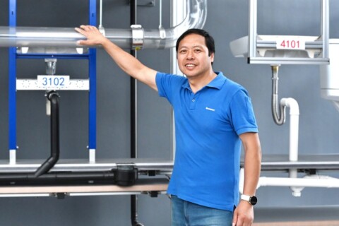 A man in a blue shirt stands beside plumbing fixtures and pipes on a display wall.