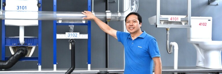 A man in a blue shirt stands beside plumbing fixtures and pipes on a display wall.
