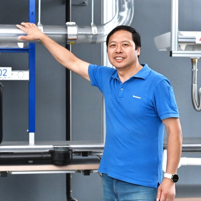 A man in a blue shirt stands beside plumbing fixtures and pipes on a display wall.