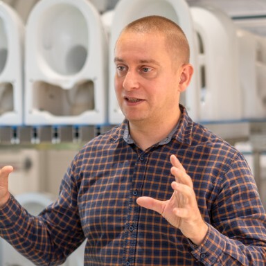Ein Mann in einem karierten Hemd spricht mit erhobenen Händen vor einer Hintergrundszene, die mehrere Toilettenkeramiken zeigt.