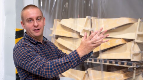 Ein Mann in karierter Bluse erklärt etwas vor einem Stapel gelber, geformter Teile, die hinter einer transparenten Folie stehen.