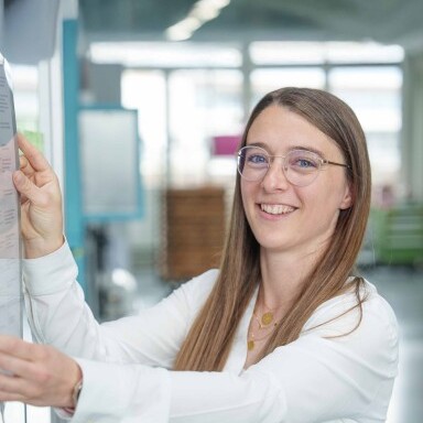 Eine junge Frau mit langen, braunen Haaren und einer weissen Bluse lächelt.