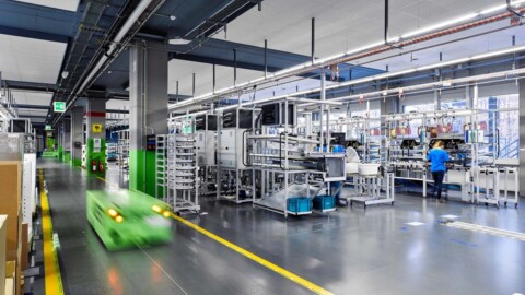 A large industrial warehouse with assembly stations, equipment, and a worker in blue, featuring a green automated cart moving along a marked path.