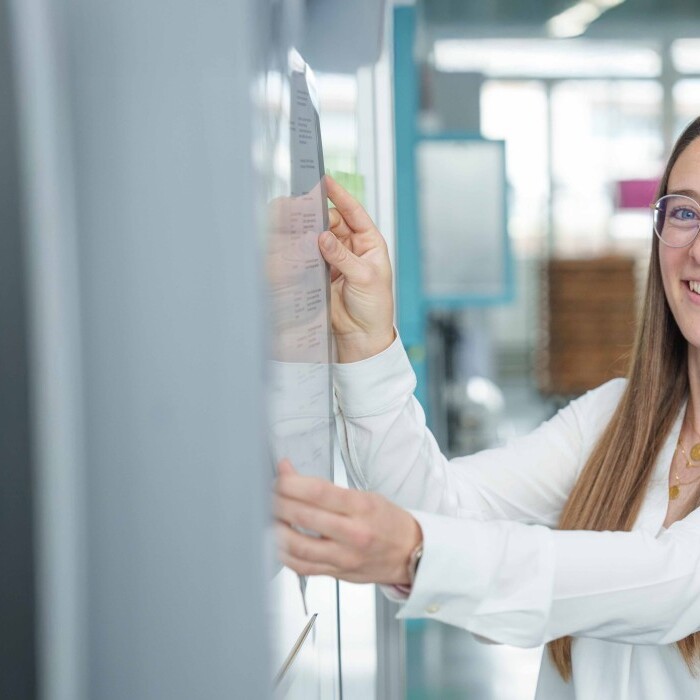 A young woman with long hair and glasses is smiling while standing in a modern environment.