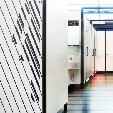 Weisse Trennwände in einem modernen Waschraum, teilweise mit Streifenmuster, mit Spiegel und Waschbecken im Hintergrund.