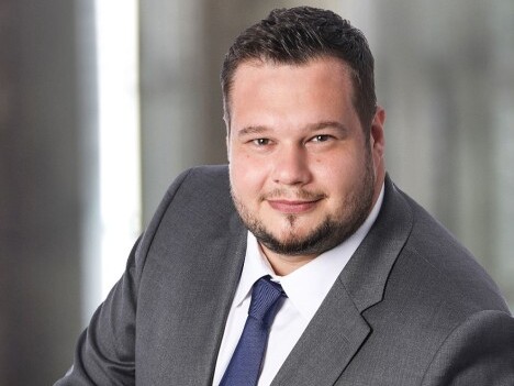 Mann in Anzug und Krawatte, lächelt freundlich in einer neutralen Umgebung mit unscharfem Hintergrund.