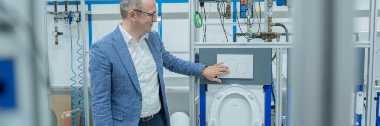 Ein Mann in einem Anzug steht vor einem technischen Gerät, das eine WC-Spülung demonstriert, und berührt das Bedienfeld.