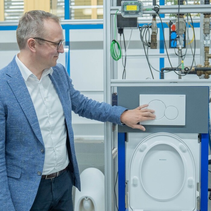 Ein Mann in einem Anzug steht vor einem technischen Gerät, das eine WC-Spülung demonstriert, und berührt das Bedienfeld.