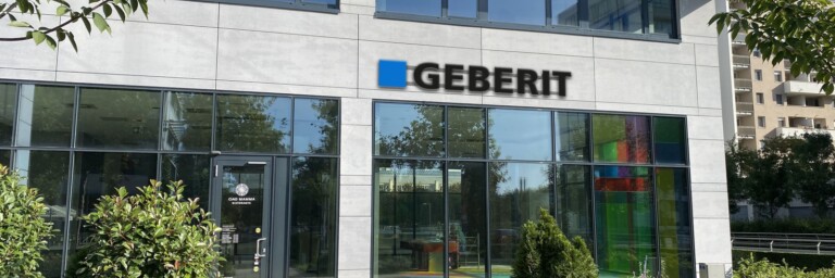 Modern glass-front building with large windows and a prominent sign displaying the name "GEBERIT," surrounded by greenery.