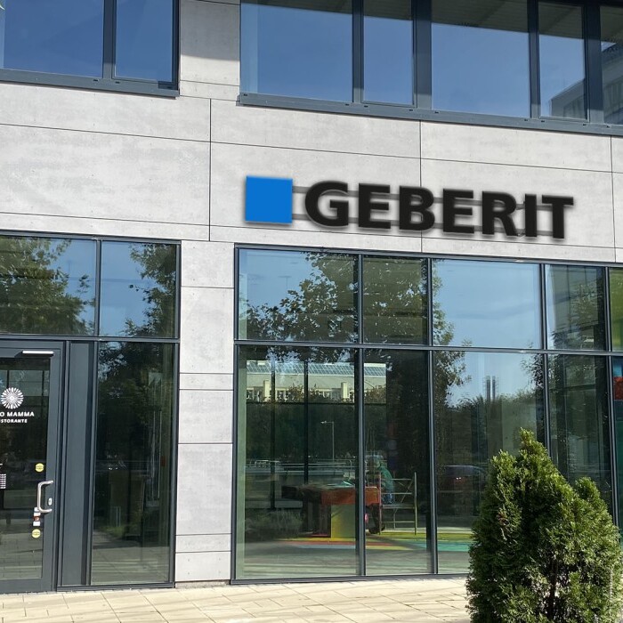 Modern glass-front building with large windows and a prominent sign displaying the name "GEBERIT," surrounded by greenery.