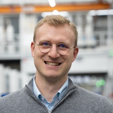 A man with short blonde hair and glasses smiles while wearing a gray sweater over a light blue shirt, set against an industrial background.