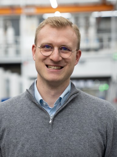 A man with short blonde hair and glasses smiles while wearing a gray sweater over a light blue shirt, set against an industrial background.