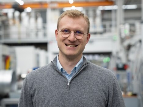 Ein lächelnder Mann mit Brille trägt einen grauen Pullover in einer industriellen Umgebung mit Maschinen im Hintergrund.