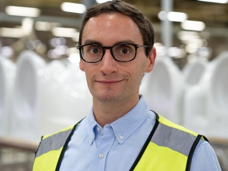 Ein Mann mit Brille trägt eine gelbe Warnweste und steht in einer Fabrik mit weissen Objekten im Hintergrund.