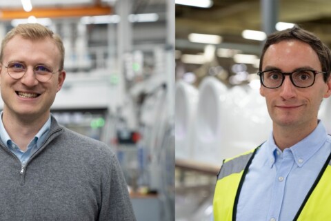 Two men are smiling in a factory setting; one wears a gray sweater, and the other wears a yellow safety vest over a light blue shirt.