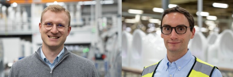 Zwei Männer stehen in einem Produktionsumfeld; der erste trägt einen grauen Pullover und der zweite eine gelbe Sicherheitsweste über einem Hemd.