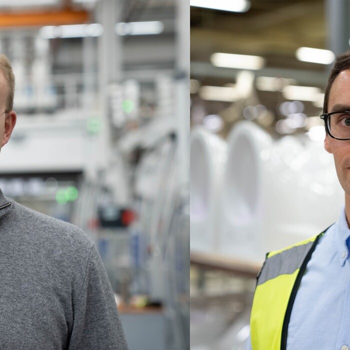 Zwei Männer stehen in einem Produktionsumfeld; der erste trägt einen grauen Pullover und der zweite eine gelbe Sicherheitsweste über einem Hemd.