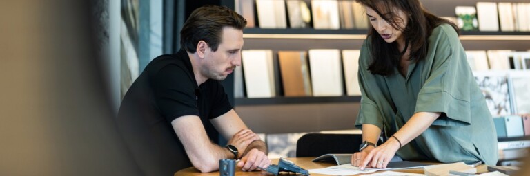 Two people discussing and analyzing documents at a table, with design samples visible in the background.