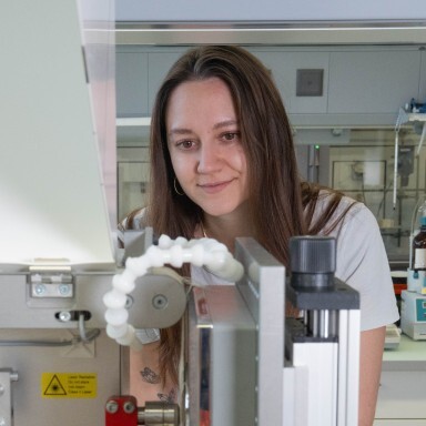 A young woman smiles while working in a laboratory, standing near a scientific instrument.