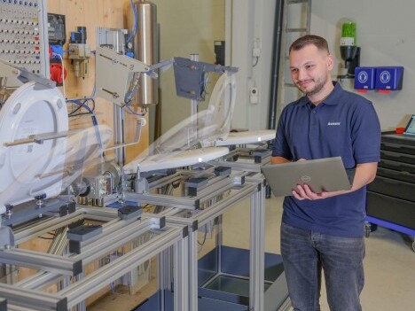 A technician operating machinery while holding a laptop in a workshop setting.