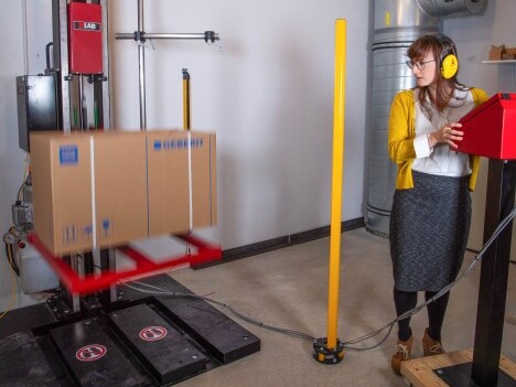 A woman in a yellow cardigan and headphones stands beside a red control panel while a box is being dropped by a machine in a warehouse setting.