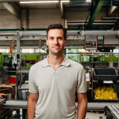 Ein Mann in einem hellen Poloshirt steht in einer modernen Produktionsumgebung mit industrieller Ausstattung im Hintergrund.