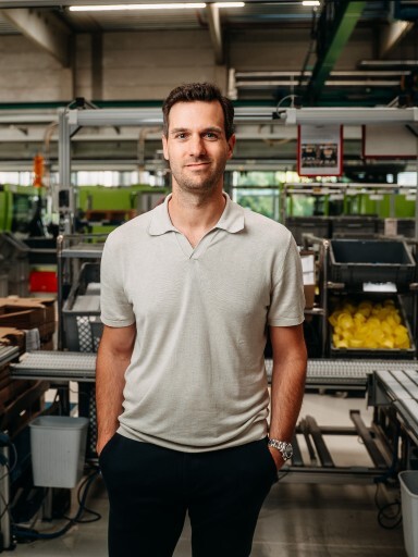 A man standing in a factory setting, wearing a light gray polo shirt and dark pants, with machinery and equipment in the background.