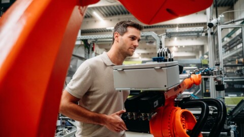 A man holding a control box near a robotic arm in a manufacturing setting.
