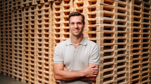 Man standing with arms crossed in front of a stack of wooden pallets.