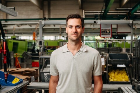 A man in a light grey polo shirt stands confidently among various industrial equipment and containers in a factory setting.