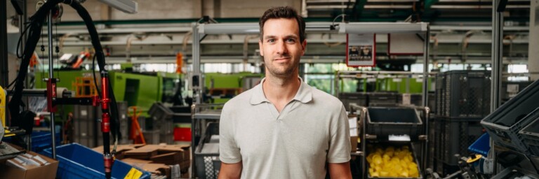 A man in a light grey polo shirt stands confidently among various industrial equipment and containers in a factory setting.
