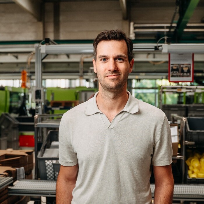 A man in a light grey polo shirt stands confidently among various industrial equipment and containers in a factory setting.