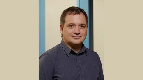 A man with short brown hair, wearing a dark blue shirt, standing in front of a light-colored wall with blue accents.