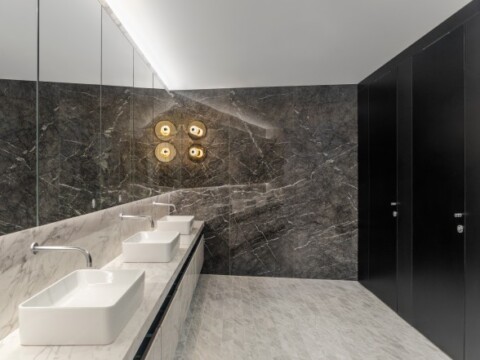 Modern restroom featuring a long marble countertop with three rectangular sinks, a large mirror, decorative wall lighting, and dark private stalls.