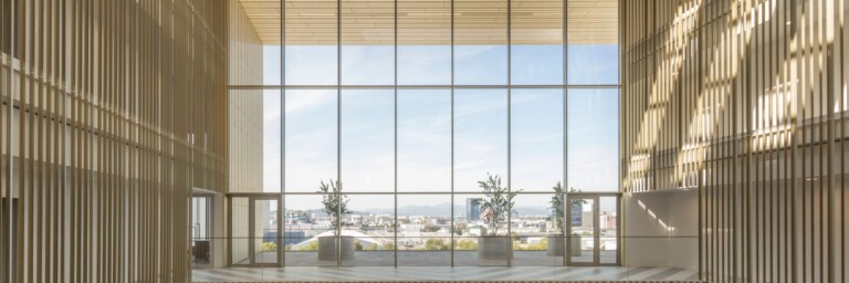 A modern interior space featuring large glass windows, wooden beams, and potted plants, with a view of the outdoors.