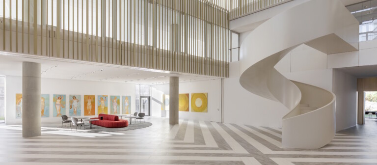A modern interior of a spacious lobby featuring a spiral staircase, gray flooring, and a red sofa surrounded by artwork.