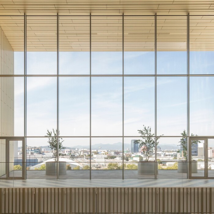 A modern interior space featuring large glass windows, wooden beams, and potted plants, with a view of the outdoors.