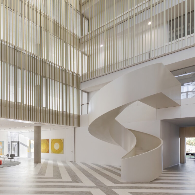 A modern interior of a spacious lobby featuring a spiral staircase, gray flooring, and a red sofa surrounded by artwork.