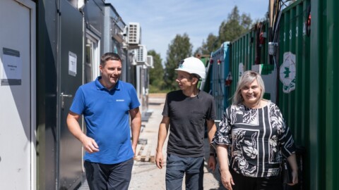 Three individuals walking together on a construction site, surrounded by shipping containers and portable buildings, under a clear blue sky.